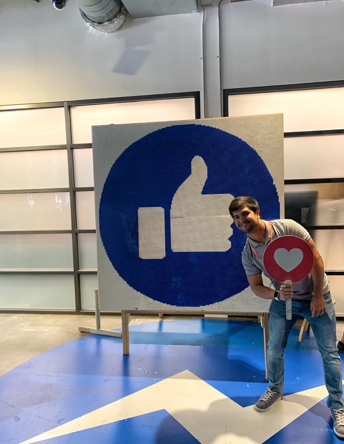 Matheus Siqueira beside a large pixel-mosaic thumbs-up, holding a round paddle with a heart on it.