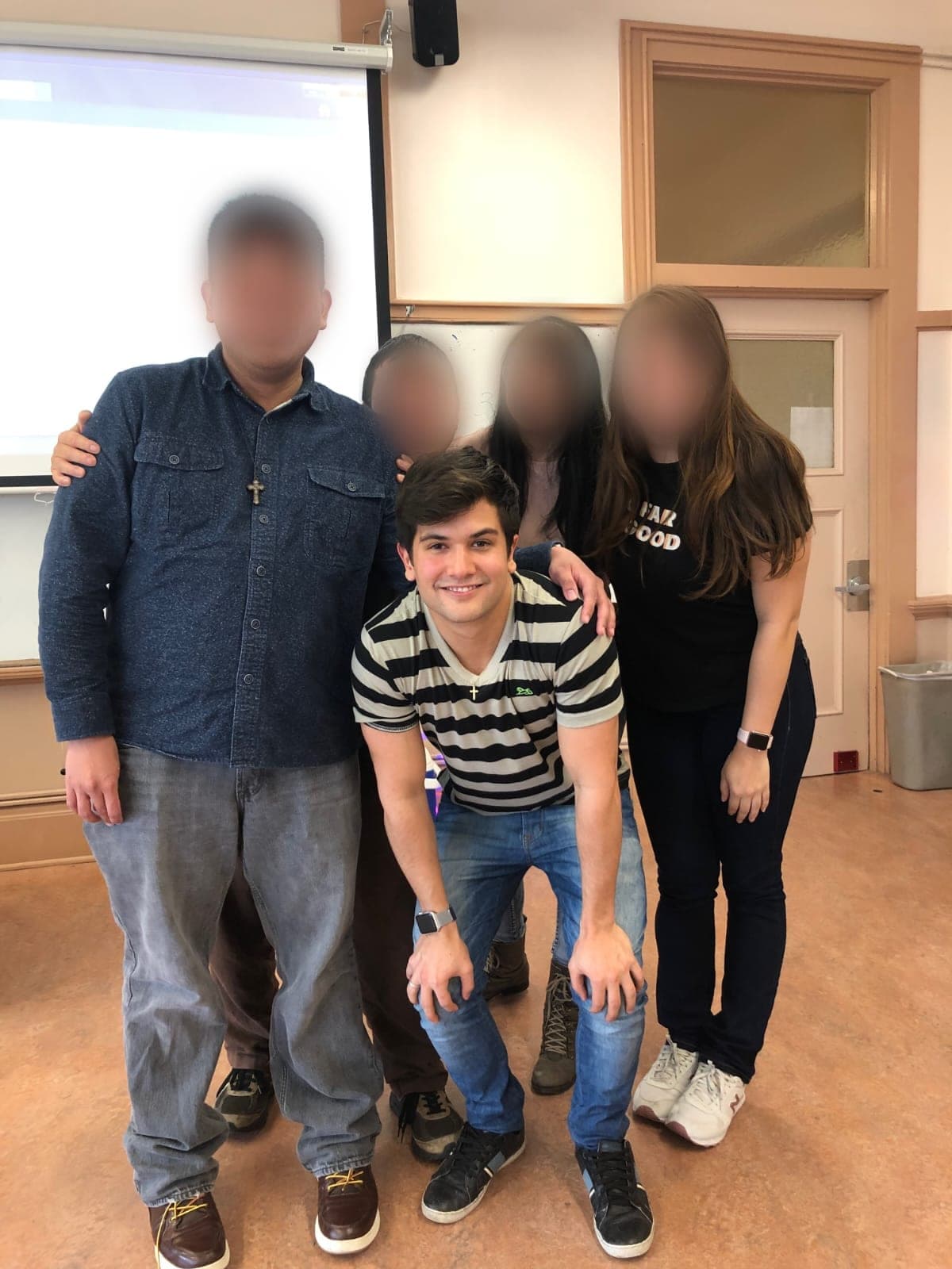Matheus Siqueira crouching in front of four people with blurred faces, in a room with a projection screen and whiteboard.